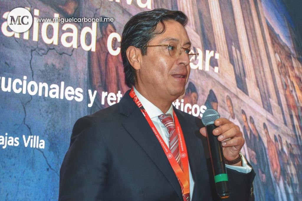 Hombre de cabello oscuro y gafas hablando en un evento, sosteniendo un micrófono. Detrás de él, hay una pantalla con texto relacionado con la calidad en la educación.