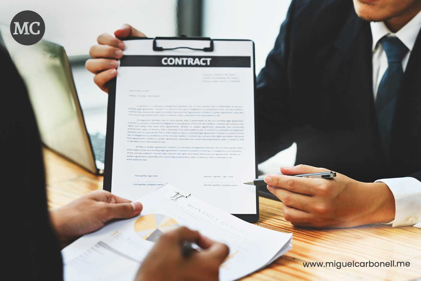 Dos personas discutiendo un contrato. Una sostiene el contrato mientras la otra toma notas con un bolígrafo, sobre una mesa con gráficos y un ordenador portátil al fondo.