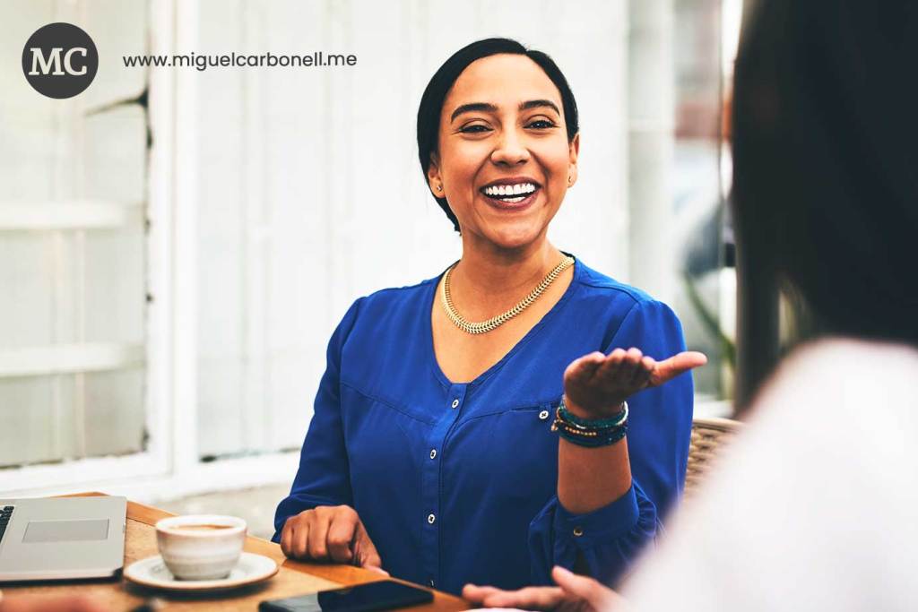 Mujer sonriendo y hablando en una cafetería, vestida con una blusa azul, con una taza de café en la mesa.