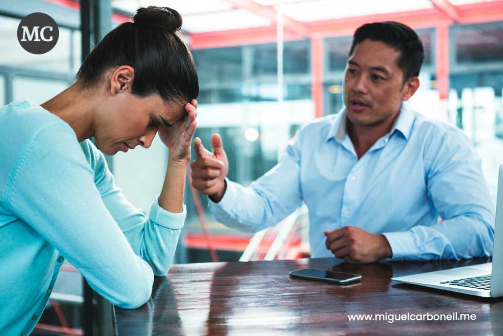 Una mujer con expresión de preocupación se sostiene la frente mientras un hombre le habla, gesticulando con las manos en una reunión de trabajo.