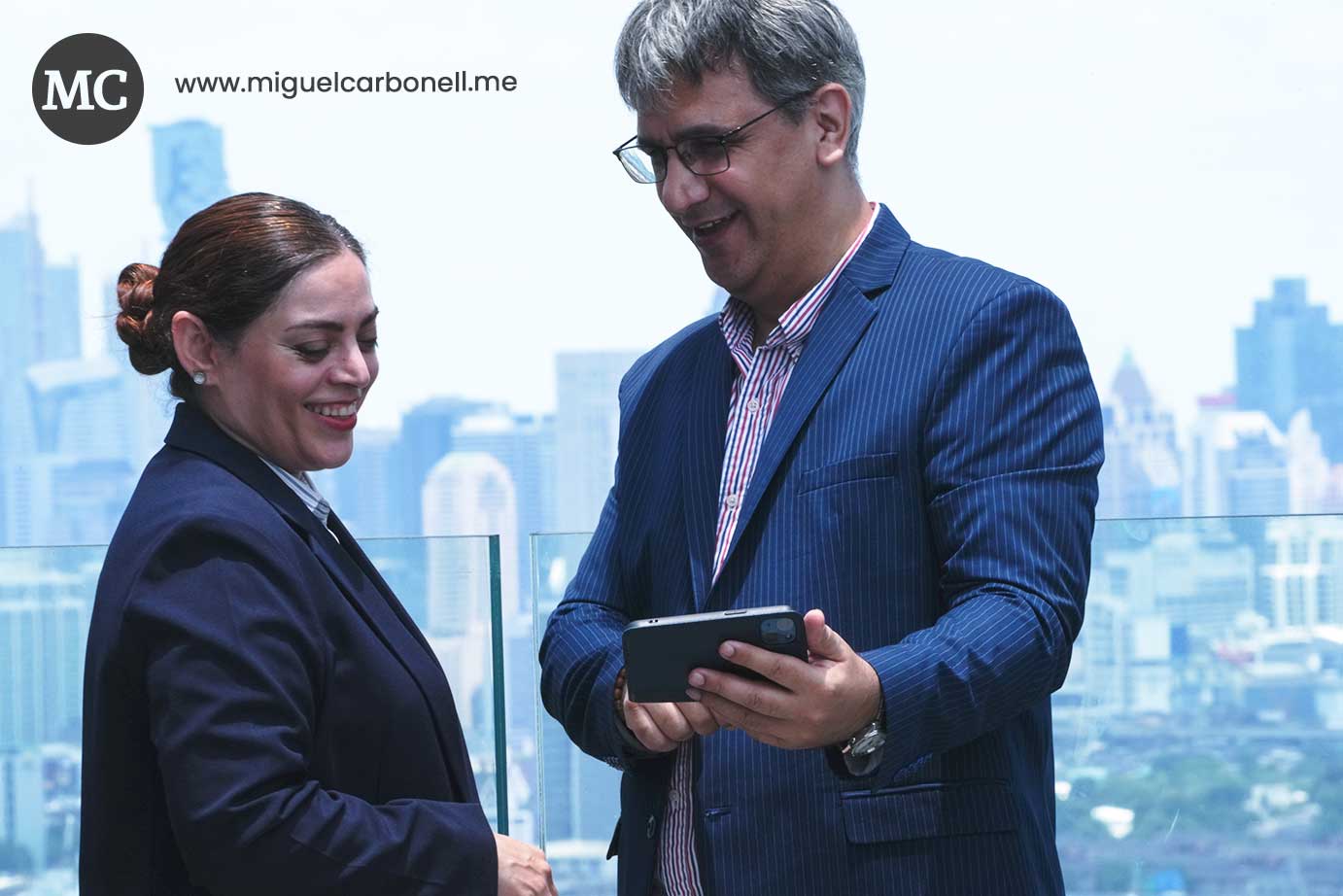 Dos personas de negocios sonríen y miran una tablet en un entorno urbano, con rascacielos de fondo.