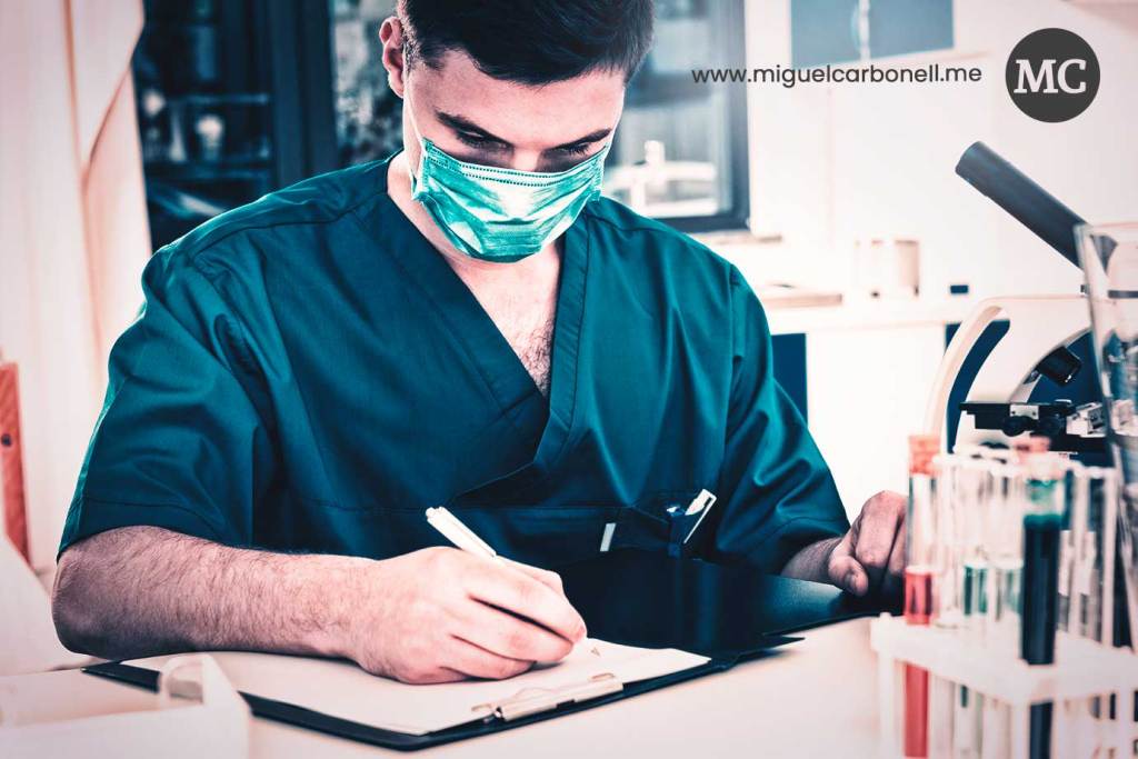 Hombre en un laboratorio médico, usando una mascarilla y camisa de scrubs, escribiendo en una hoja en una mesa con tubos de ensayo y un microscopio cerca.