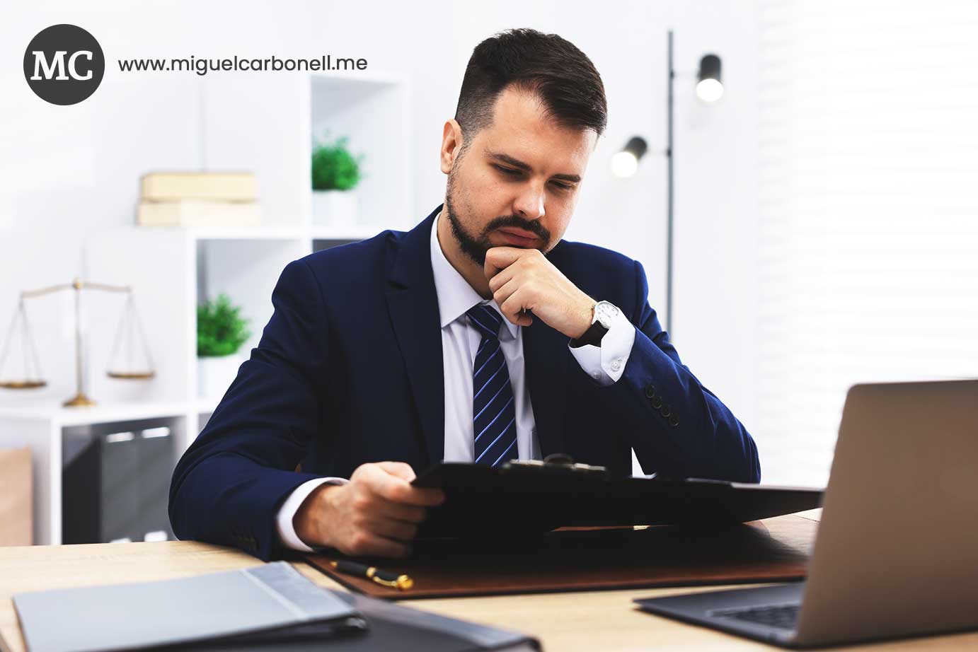 Hombre de negocios vestido de traje azul, sentado en un escritorio con una carpeta en las manos, pensativo en un entorno de oficina moderna.