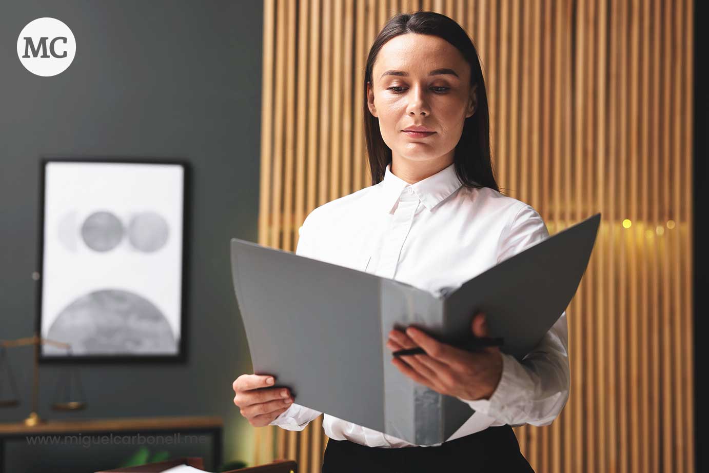 Mujer de pie sosteniendo un folder, vestida con camisa blanca, en un entorno de oficina elegante con paredes de madera.